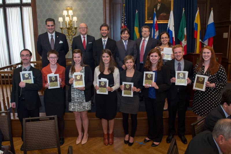 Group of award recipients and officials posing for a photo indoors; the front row holds certificates or plaques, with international flags displayed in the background.